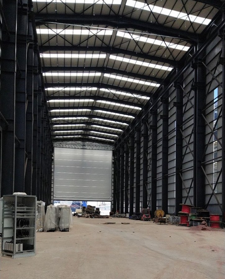 Interior of a large covered fabrication hall with overhead cranes and a high bay door.