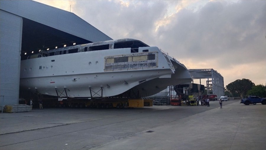 A large white hull being moved out of a fabrication shed on a shipyard transporter.